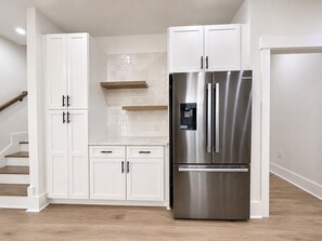 A modern kitchen with white cabinetry, a stainless steel refrigerator, and a wooden staircase. - 5 Bedroom, Private Pool, Elevator - New Construction - Why Knot (Surfside Beach)