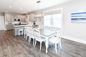 A modern kitchen with white cabinetry, a central island, and a dining area with white chairs and a glass table. - 5 Bedroom, Private Pool, Elevator - New Construction - Why Knot (Surfside Beach)