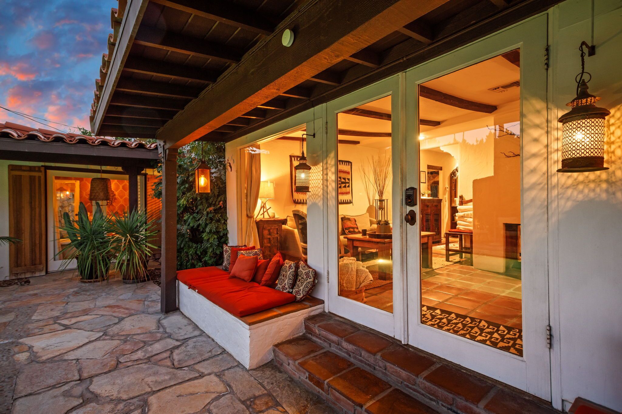 A cozy outdoor seating area with a stone floor, a red cushioned bench, and a view into the interior of a home.
