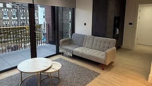 Ein modernes Wohnzimmer mit grauem Sofa, einem kleinen runden Tisch und einer großen Glasschiebetür, die auf einen Balkon im Freien führt.