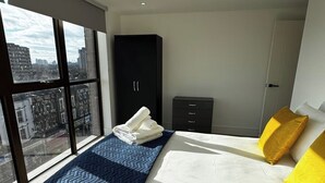 Ein modernes Schlafzimmer mit einem großen Bett, einem Kleiderschrank, einer kleinen Kommode und Blick auf die Stadt durch große Fenster.