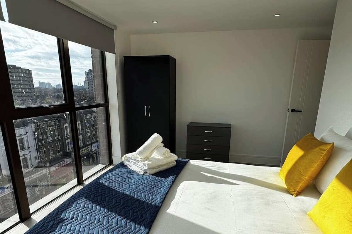 A modern bedroom with a large bed, a wardrobe, a small cabinet, and a view of the city through large windows.
