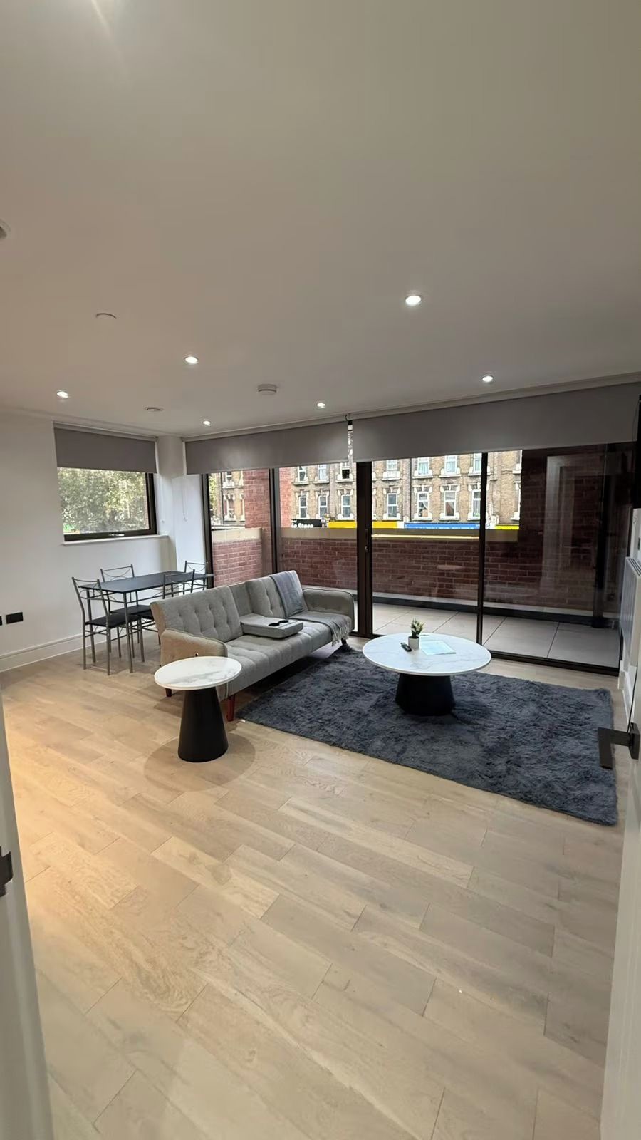 A modern living room with a sectional sofa, a round coffee table, and a dining area with a table and chairs. Large windows offer a view of the city.