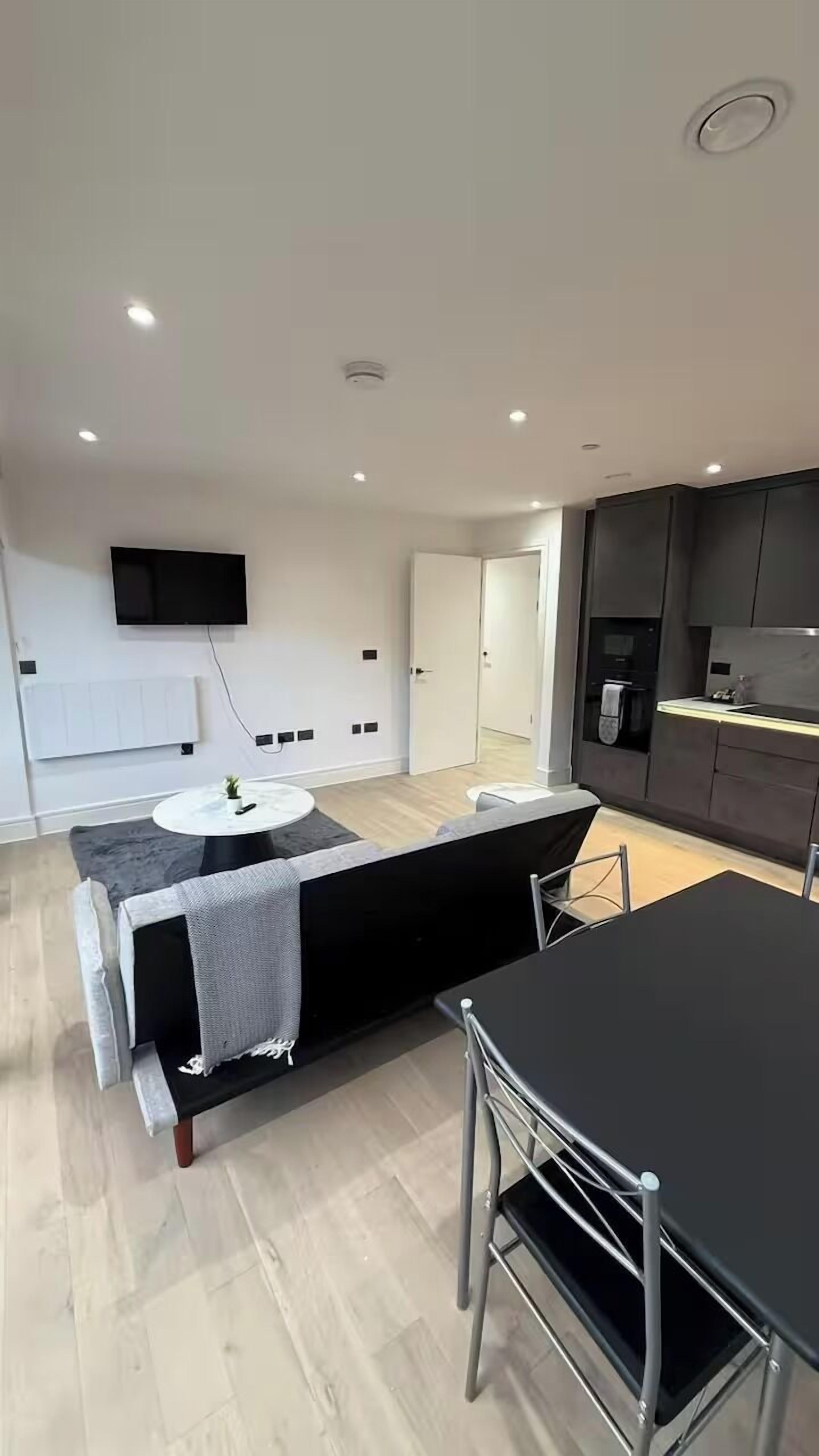 A modern living room with a black sofa, a round coffee table, and a TV mounted on the wall. There is a kitchen area with a stove and cabinets.