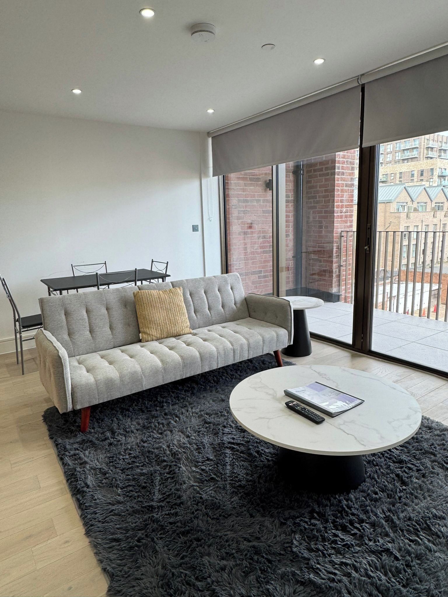 A modern living room with a grey sofa, a small round coffee table, and a view of a brick building through large windows.