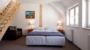 A bedroom with a wooden staircase, a bed with white and patterned bedding, two bedside lamps, a nightstand, a painting on the wall, and a window with blinds. - ZORZA (Sztutowo)