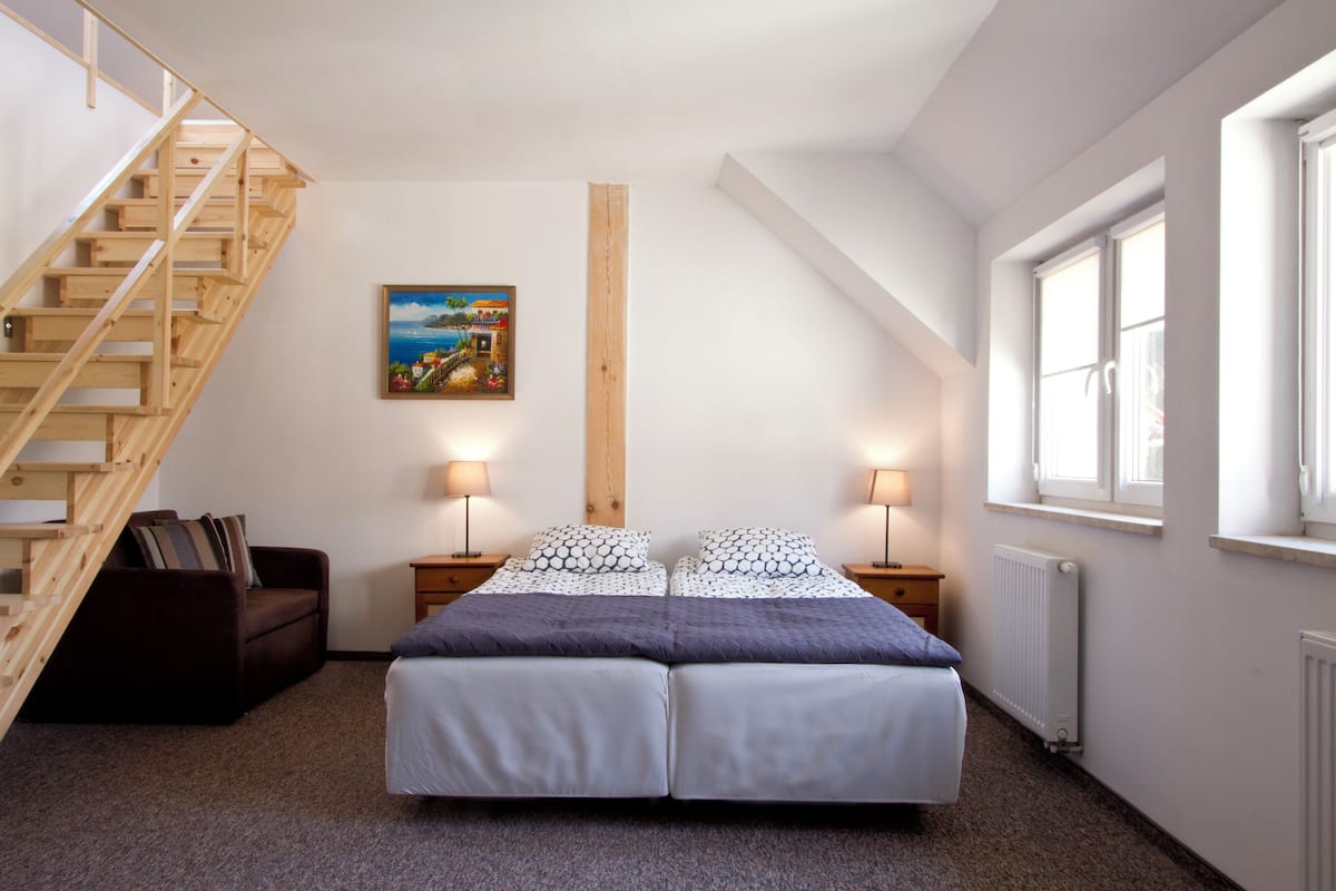 A bedroom with a wooden staircase, a bed with white and patterned bedding, two bedside lamps, a nightstand, a painting on the wall, and a window with blinds.