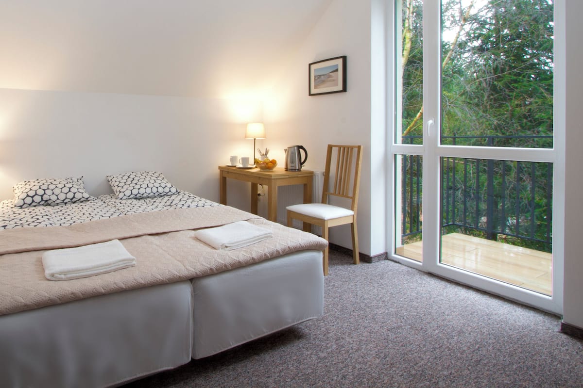 A bedroom with a large bed, two bedside tables, a chair, a small table with a lamp, and a view of greenery through a window.