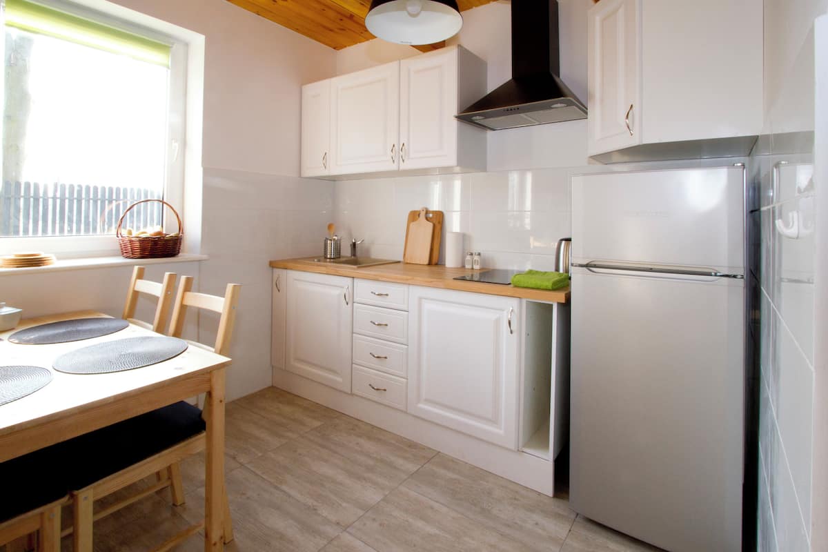 A kitchen with white cabinets, a stainless steel refrigerator, a wooden dining table with chairs, and a window with a basket on the sill.