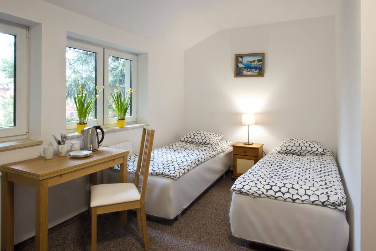 A small, tidy room with two single beds, a wooden desk, a chair, a small table with a kettle, and a lamp.