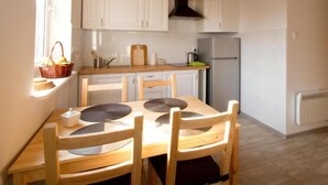 A small kitchen with white cabinets, a wooden table with four chairs, a window with a basket of fruit, and a refrigerator. - ZORZA (Sztutowo)