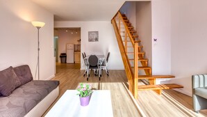 A living room with a sofa, a coffee table with a plant, a dining table with chairs, and a wooden staircase. - Gîte Acacia - Domaine de l'Enclos (Mireval)