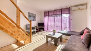 A living room with a wooden staircase, a sofa, a coffee table, a TV, and a window with purple curtains. - Gîte Acacia - Domaine de l'Enclos (Mireval)