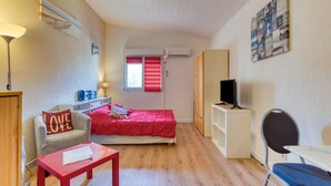 Habitación pequeña con cama, escritorio, silla, lámpara y televisor.