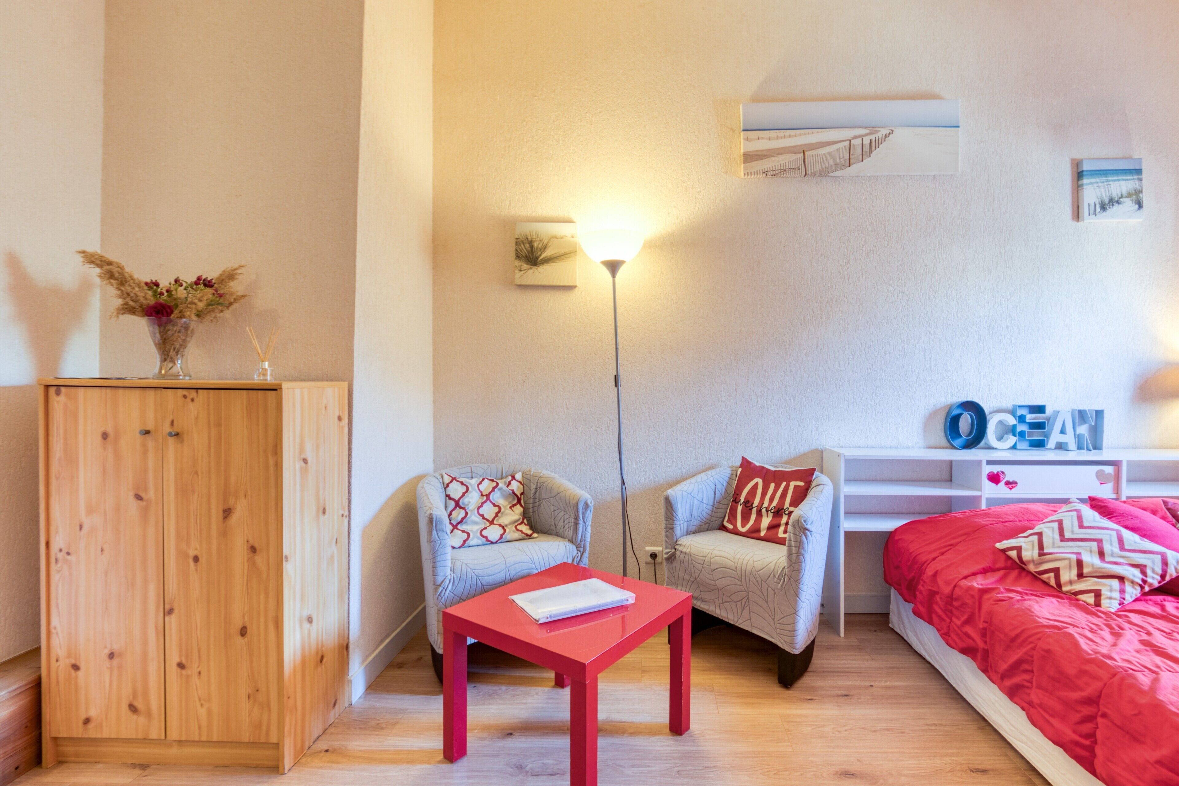 Une chambre avec une armoire en bois, une table rouge, deux fauteuils, un lit avec une literie rouge, une étagère murale ornée d’objets décoratifs et une lampe fixée au mur.