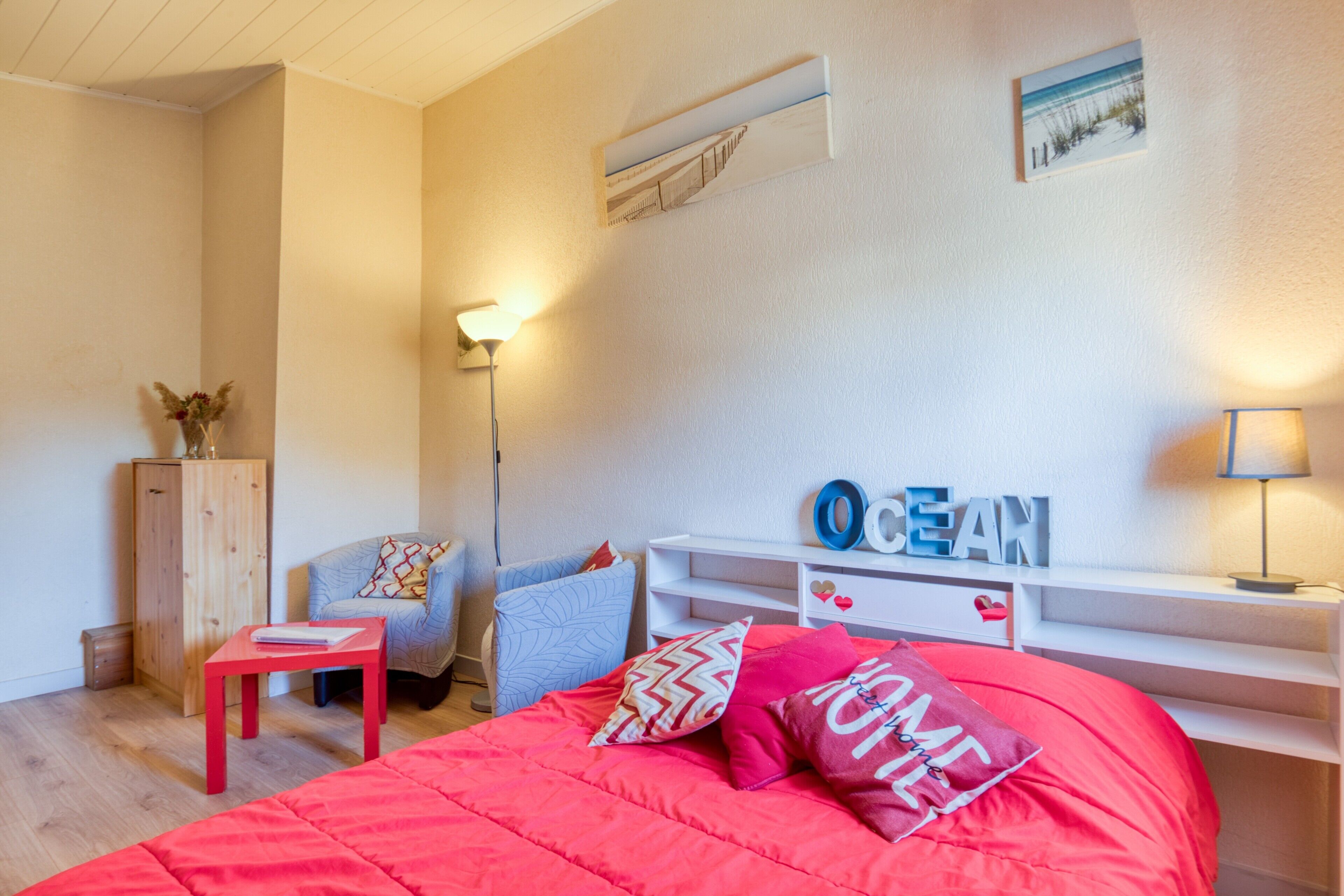 A bedroom with a red bed, a wooden cabinet, a chair, a desk with 'OCEAN' on it, and a wall-mounted shelf.