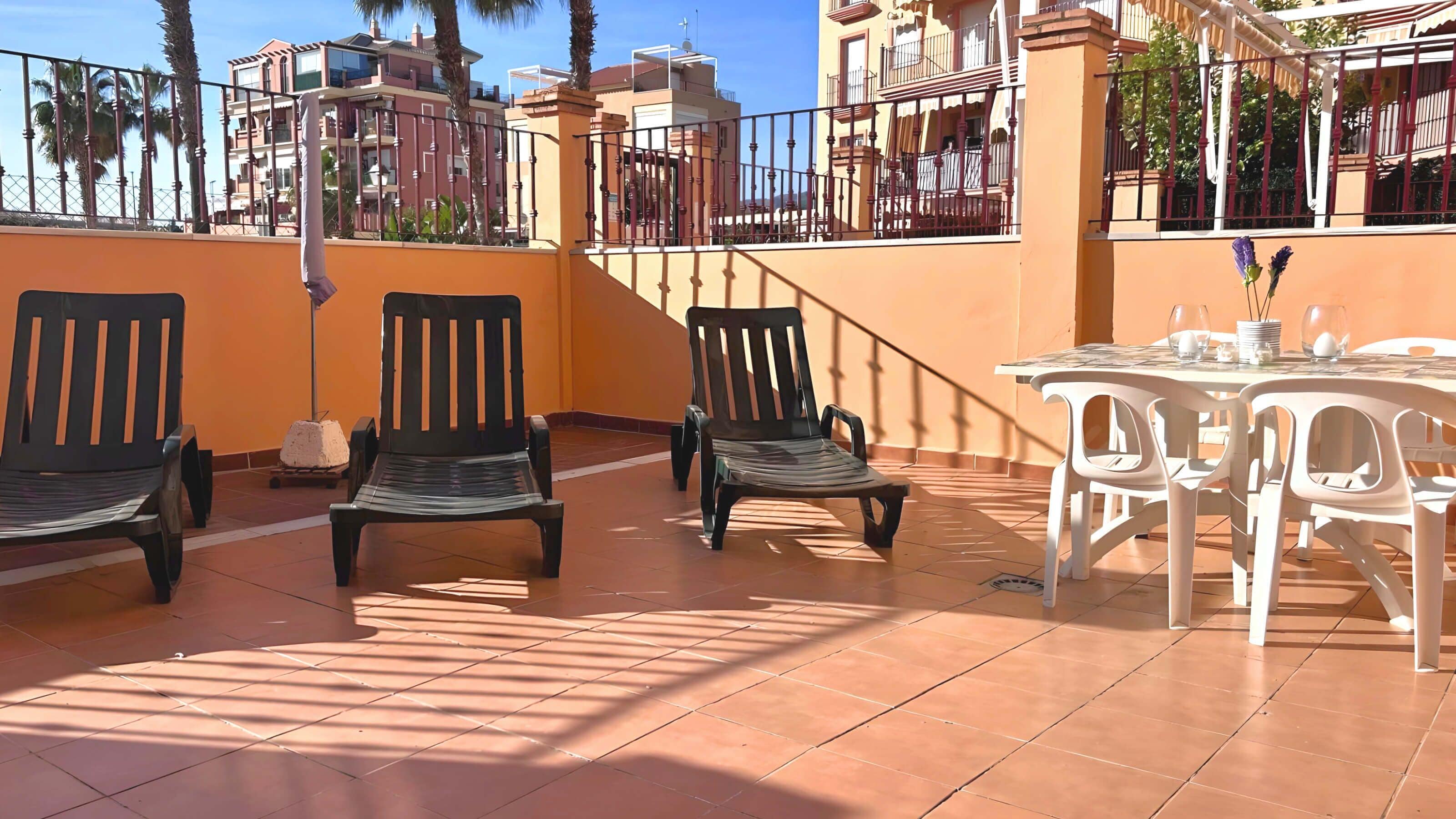 Eine Terrasse im Freien mit drei schwarzen Stühlen und einem weißen Tisch, umgeben von orangefarbenen Wänden und einem gefliesten Boden.