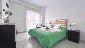 Un dormitorio con colcha verde, cabecero de madera, mesita de noche con lámpara y ventana con cortinas traslúcidas.
