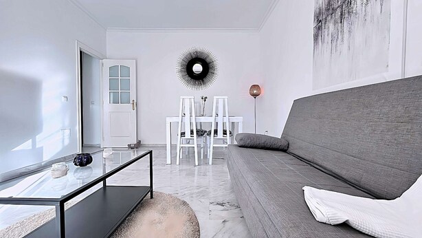 Una sala de estar moderna con un sofá gris, una mesa de centro de vidrio, una mesa de comedor blanca con sillas y una lámpara circular fijada a la pared.