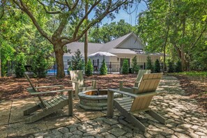 A stone patio with Adirondack chairs and a fire pit, surrounded by trees and a house in the background.