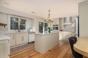 A modern kitchen with a central island, wooden flooring, and a view of trees through large windows. - Abalone Alley 04 - Easy Breezy  - Private Pool (Isle Of Palms)