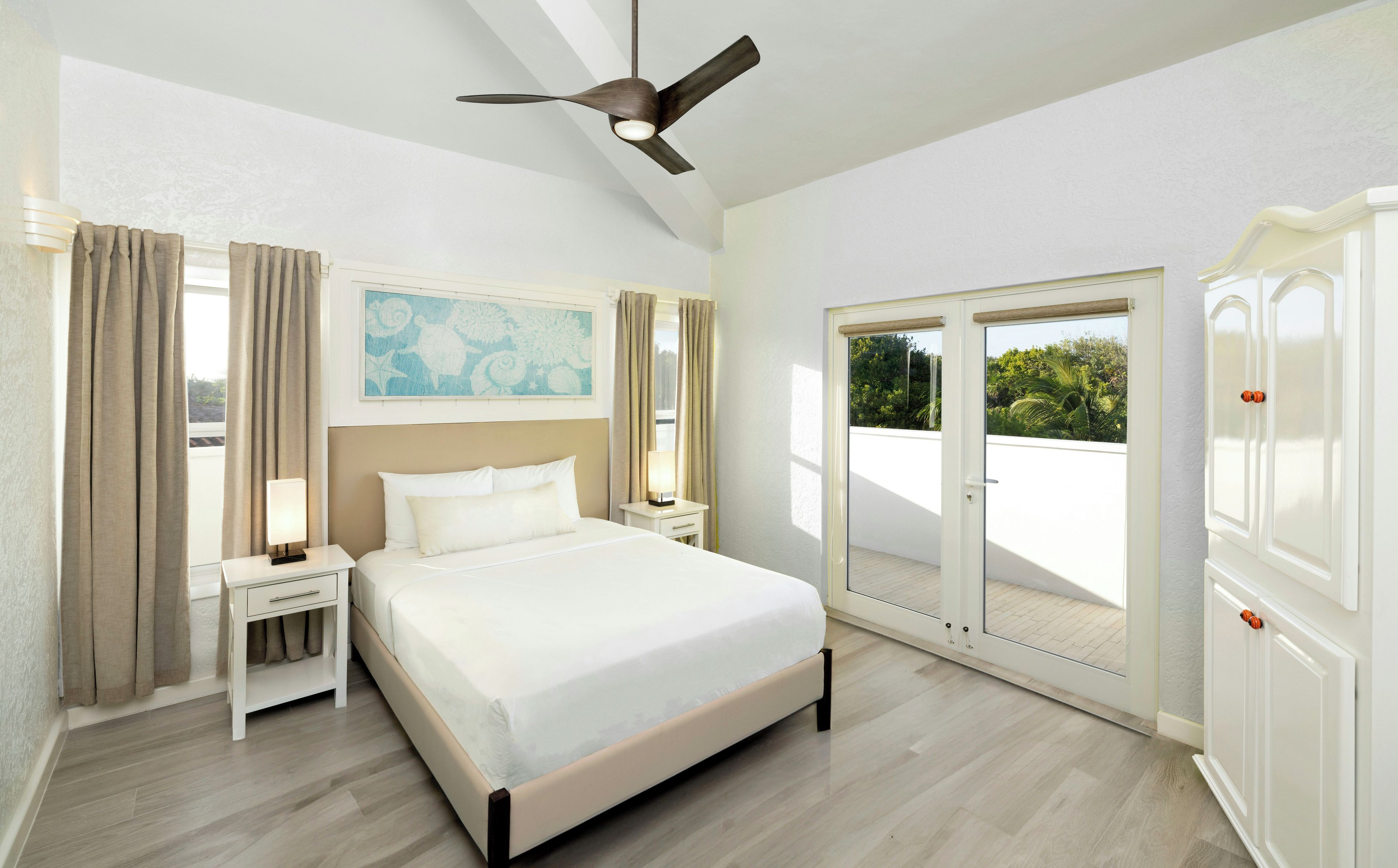 A hotel room with a bed, nightstands, a ceiling fan, a painting, and a sliding glass door to the outside.