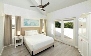Una habitación de hotel con cama, mesitas de noche, ventilador de techo, un cuadro y una puerta corrediza de vidrio al exterior.