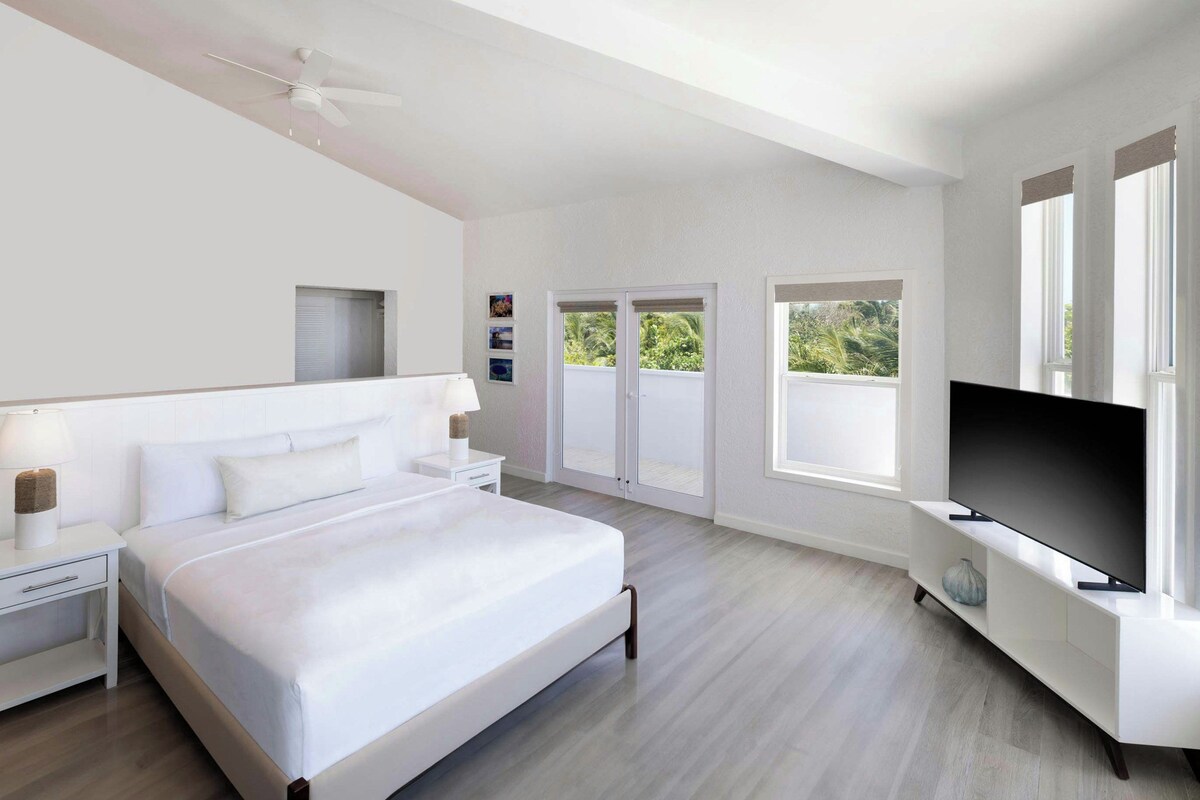 A modern bedroom with a bed, bedside tables, a ceiling fan, a TV, and a balcony with sliding doors.