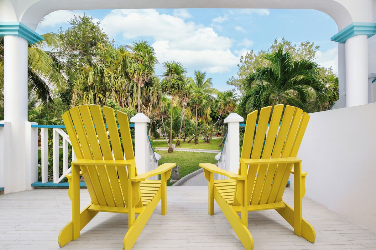 Two yellow Adirondack chairs facing a lush garden with palm trees and a clear sky.