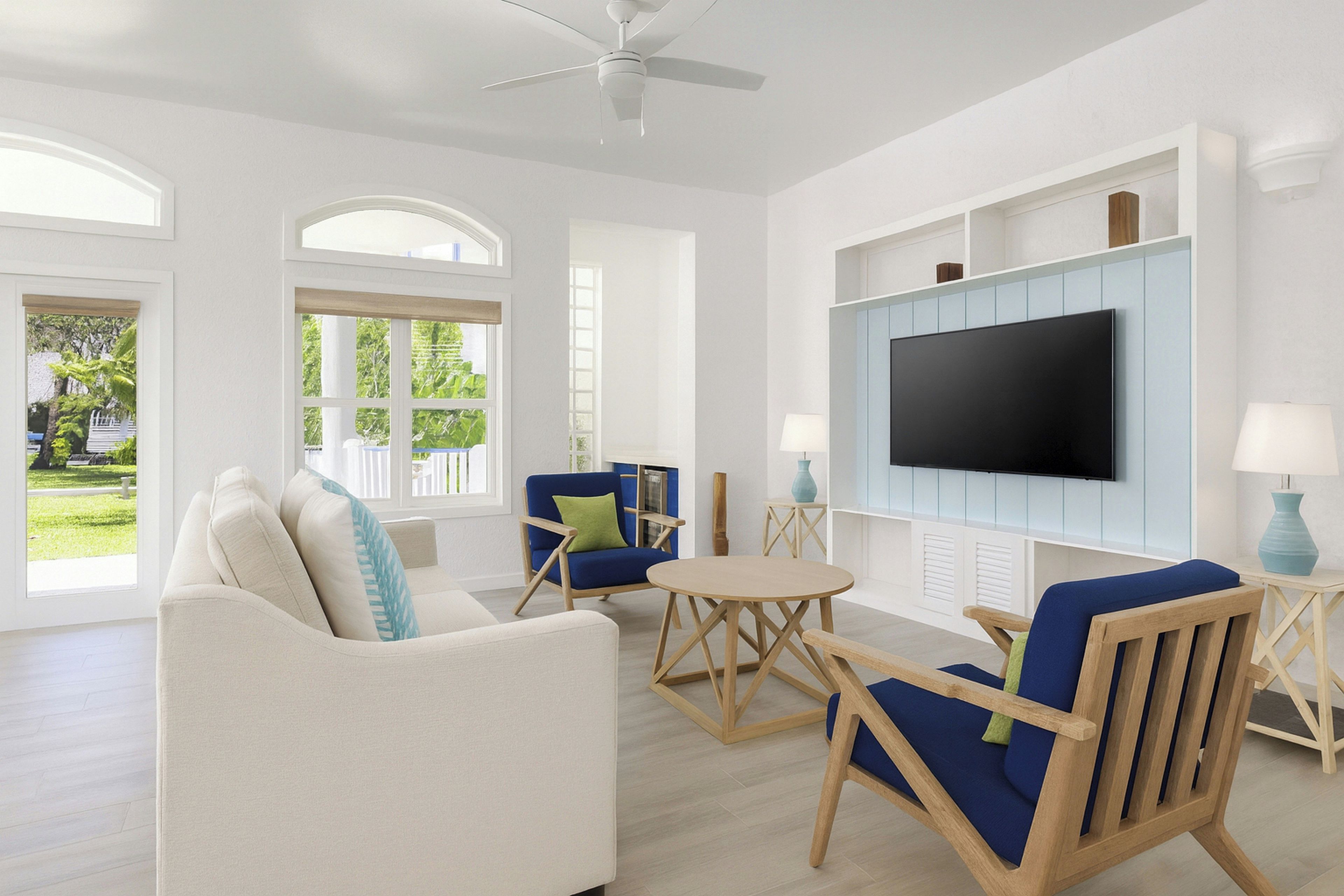A spacious living room with a white sofa, blue armchairs, a coffee table, and a flat-screen TV mounted on the wall.