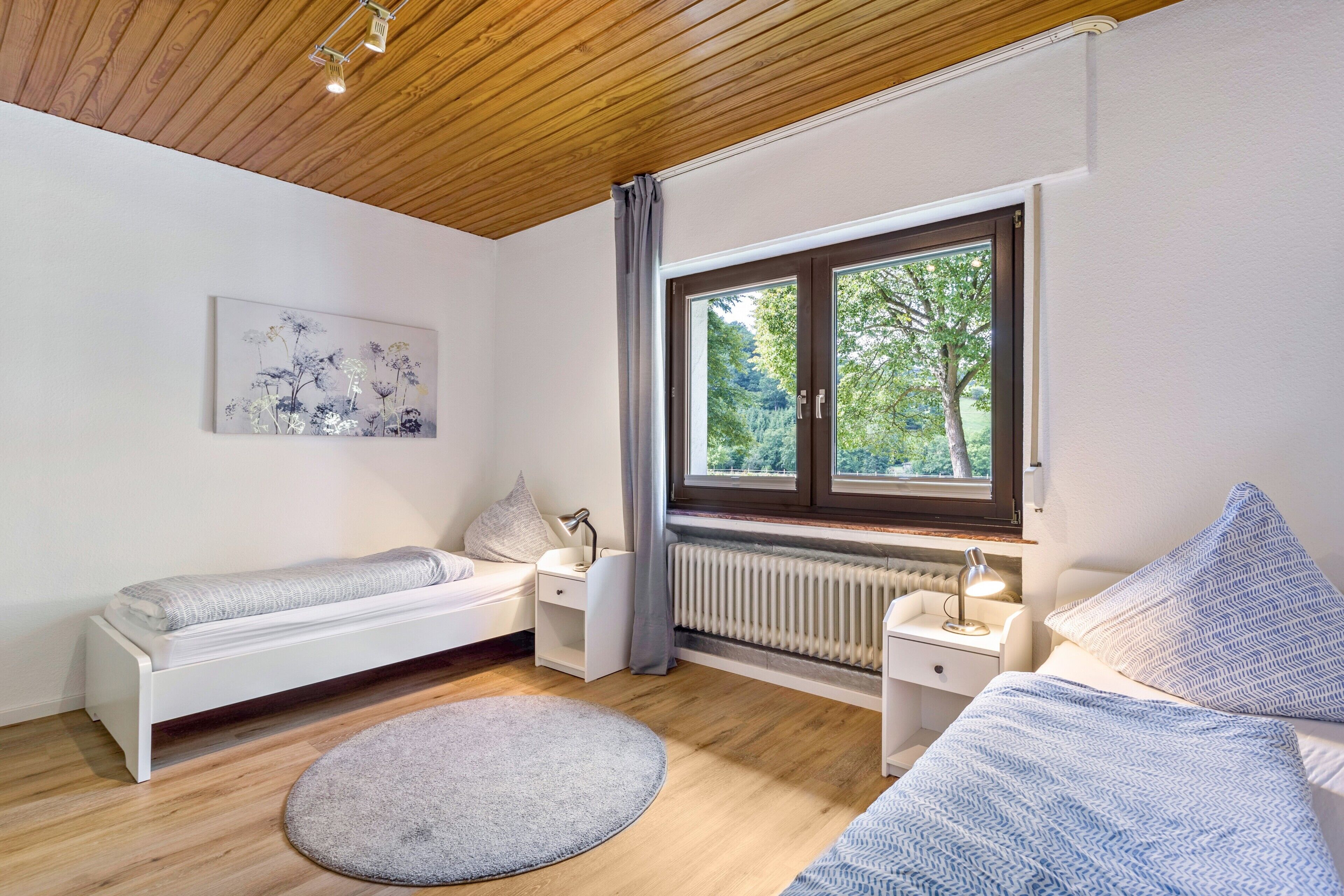Ein Schlafzimmer mit Holzbalkendecke, einem Fenster mit Blick auf Grünflächen, einem weißen Bett, einem Nachttisch mit Lampe und einer Heizung.
