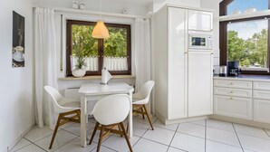 Uma cozinha moderna com armários brancos, uma mesa de jantar com cadeiras, um lustre e vista para o verde lá fora.
