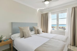 Ein Doppelbett mit Kopfteil, zwei Nachttische, eine Topfpflanze und Meerblick durch ein mit Vorhängen versehenes Fenster.