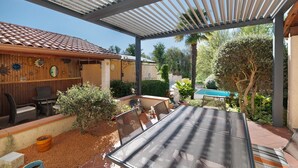 Eine überdachte Terrasse mit Tisch und Stühlen, ein Holzzaun und ein Pool im Hintergrund.