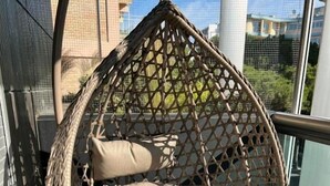 En hängande fåtölj i rotting med kuddar, placerad på en balkong med ett metallräcke och utsikt över byggnader.