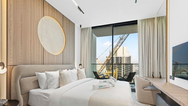 A modern hotel room with a large bed, a desk, and a view of the cityscape. - Minimalist Studio in Binghatti Azure (Jumeirah Village Circle (JVC))