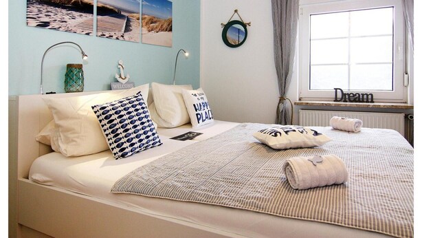 Un letto rifatto con una coperta a righe, un cuscino con la scritta “HAPPY PLACE” e un cartello con la scritta “WELCOME”. Due lampade a parete, uno specchio rotondo e una finestra con tende.
