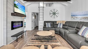 A modern living room with a sectional sofa, a flat-screen TV, a fireplace, and a large window overlooking a beach. - Rocking Sunset Beach House (Treasure Island)