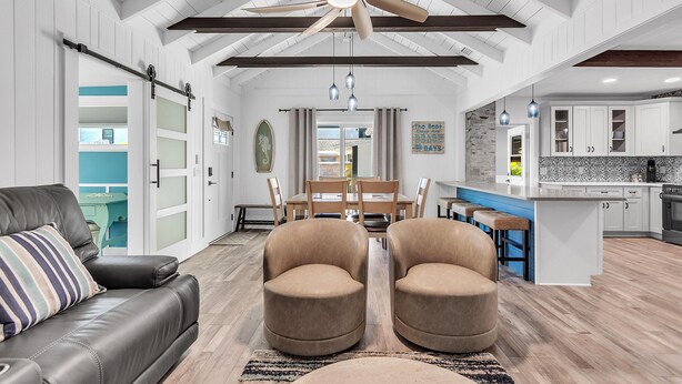 A spacious living room with a ceiling fan, a dining area with a wooden table and chairs, and a kitchen with white cabinets and a blue countertop. - Rocking Sunset Beach House (Treasure Island)