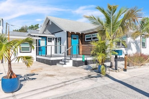 A modern two-story house with a blue door, a staircase leading to the front porch, and a small garden with palm trees. - Rocking Sunset Beach House (Treasure Island)