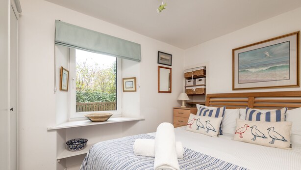 Quarto com cama, janela com vista para vegetação, mesa de cabeceira com abajur e um quadro na parede.