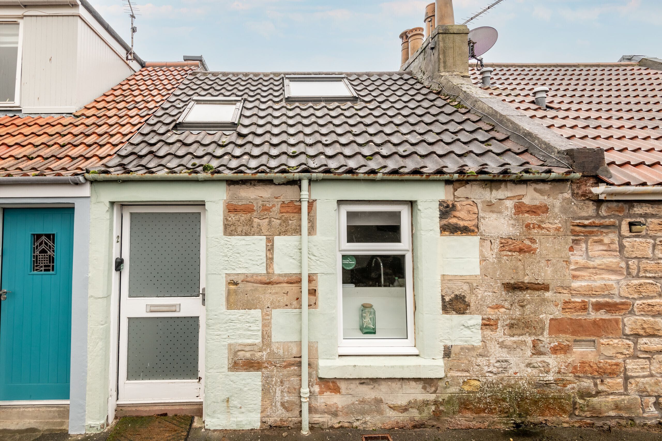 Une maison en pierre à deux étages, avec une porte de couleur turquoise, une fenêtre à cadre blanc et une antenne parabolique sur le toit.