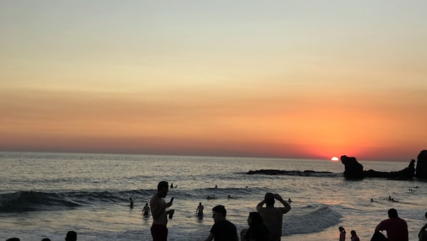 Cena de praia com pessoas nadando e permanecendo na água durante o pôr do sol.