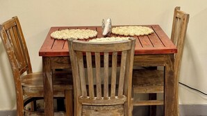 Uma mesa de jantar com quatro cadeiras de madeira, uma televisão de tela plana fixada na parede acima e um jogo americano tecido sobre a mesa.