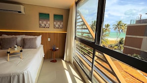 Habitación de hotel con cama, una mesita con taza y plato, una planta decorativa y un amplio ventanal con vistas a una playa y palmeras.