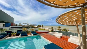 Un área de piscina con sillones de descanso y un jacuzzi, con vista a una playa con palmeras y un cielo despejado.