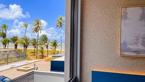 Vista desde una ventana que da a una playa con palmeras, cielo despejado y un edificio con una combinación de colores azul y beige.
