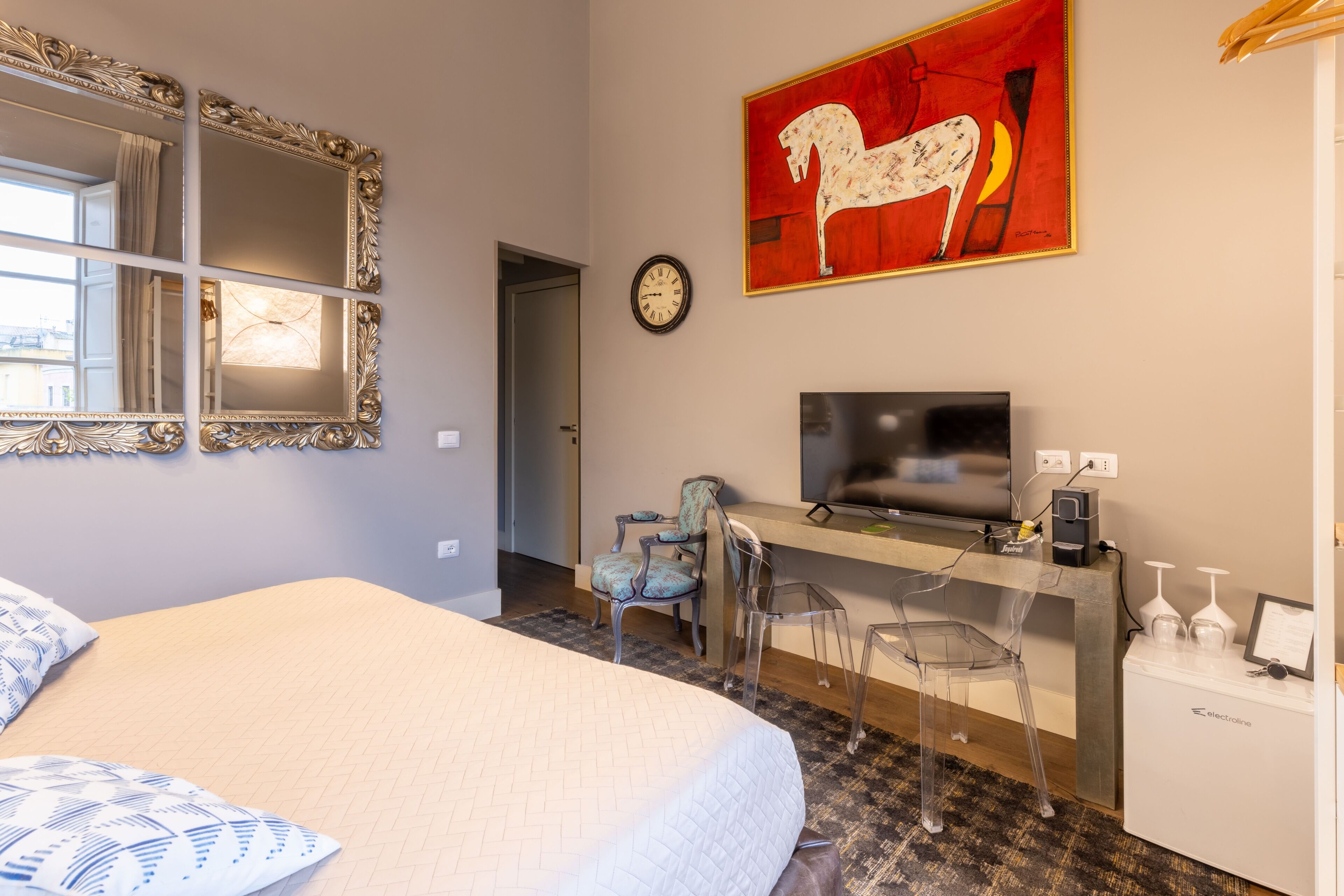 A hotel room with a bed, a desk with a TV, a clock, and a painting on the wall.