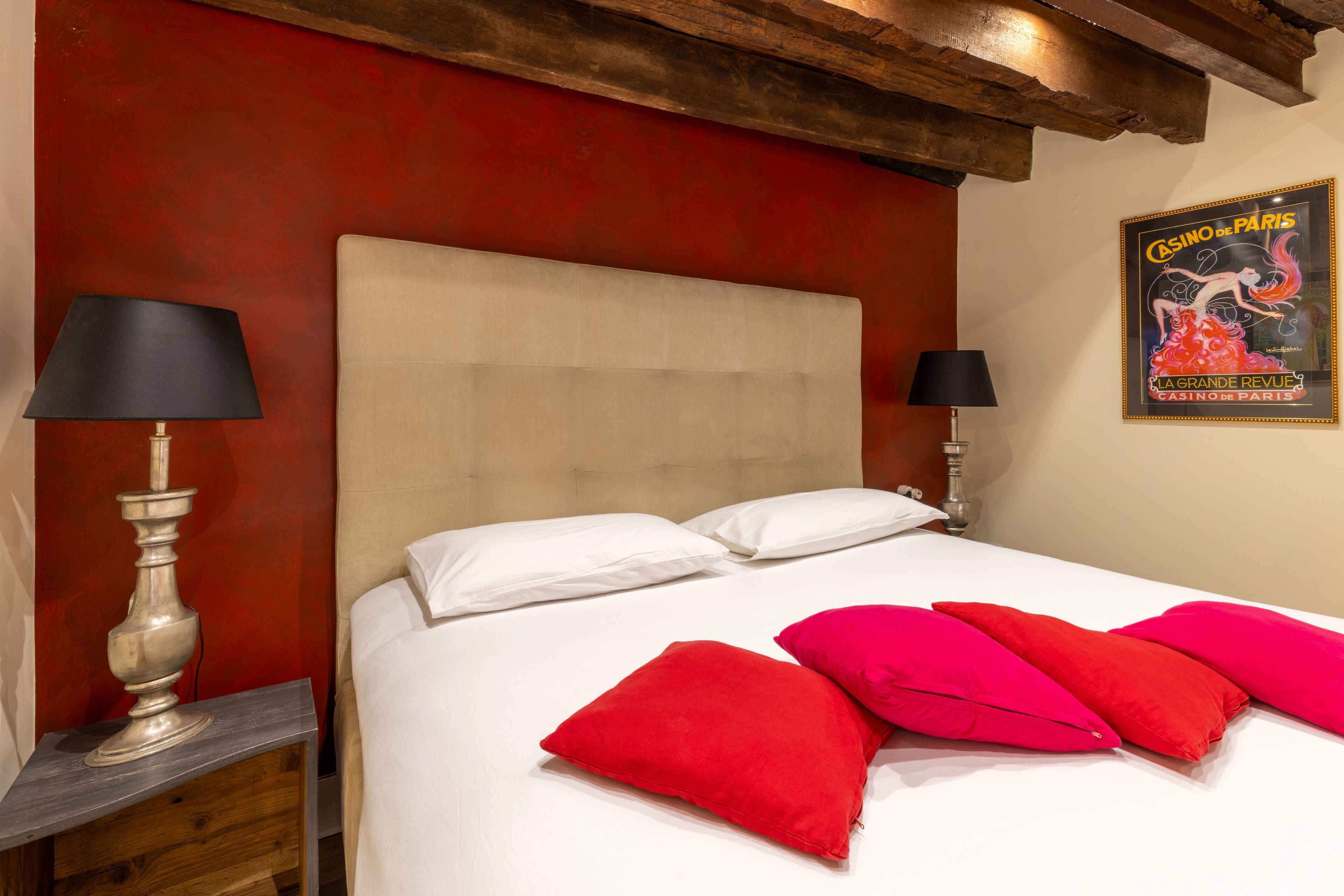 Een bed met witte en rode kussens, een houten nachtkastje en een lamp.