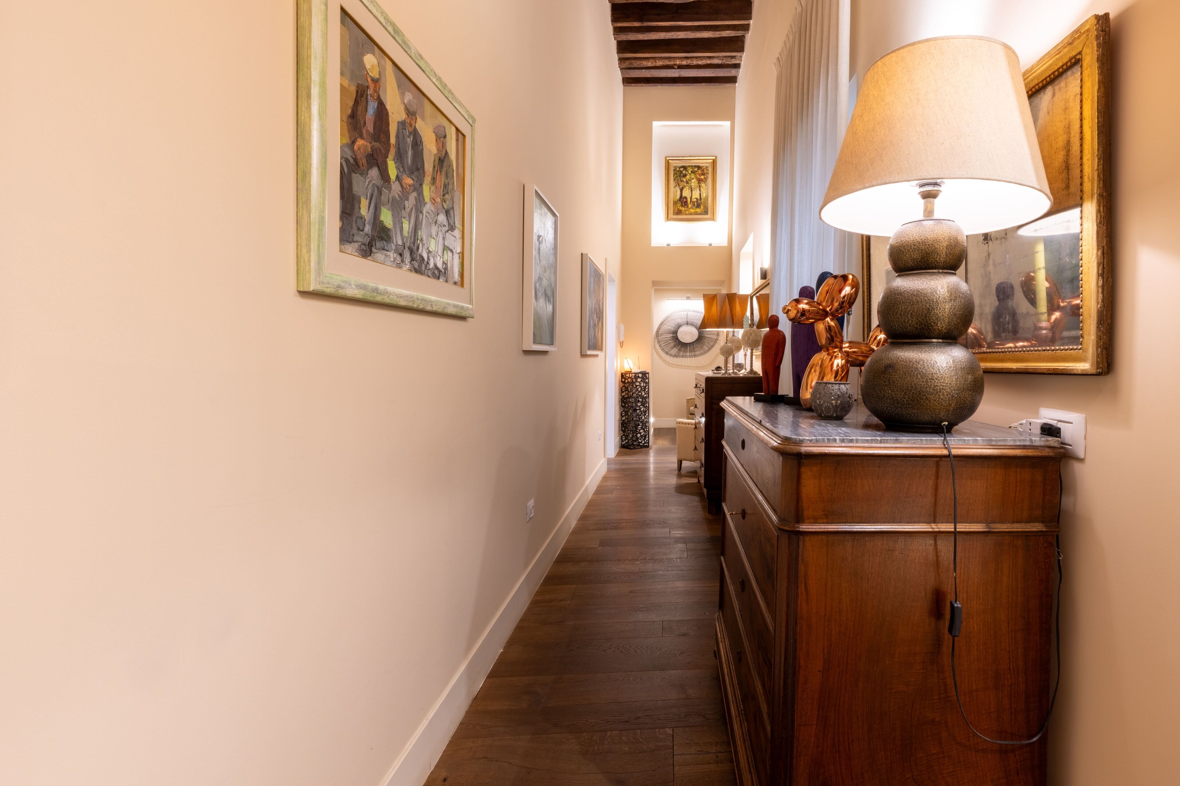 A hallway with wooden flooring, a wooden cabinet with a lamp, and framed artwork on the walls.